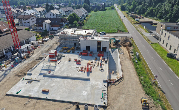 Luftaufnahme des Neubaus mit grosser Bodenplatte und teilweise errichteten Betonwänden entlang der Hauptstrasse.