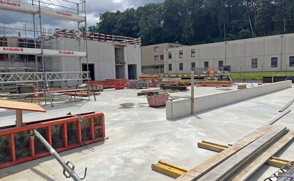 Baustelle mit Betonboden, niedrigen Betonwänden, Schalungselementen und Baugerüst unter bewölktem Himmel.